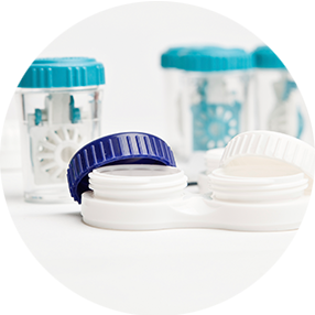 Nahaufnahme eines weißen Kontaktlinsenbehälters mit blauen und weißen Deckeln im Vordergrund und zwei klare Kontaktlinsenreinigungsbehälter mit blauen Deckeln im Hintergrund auf einer weißen Fläche.